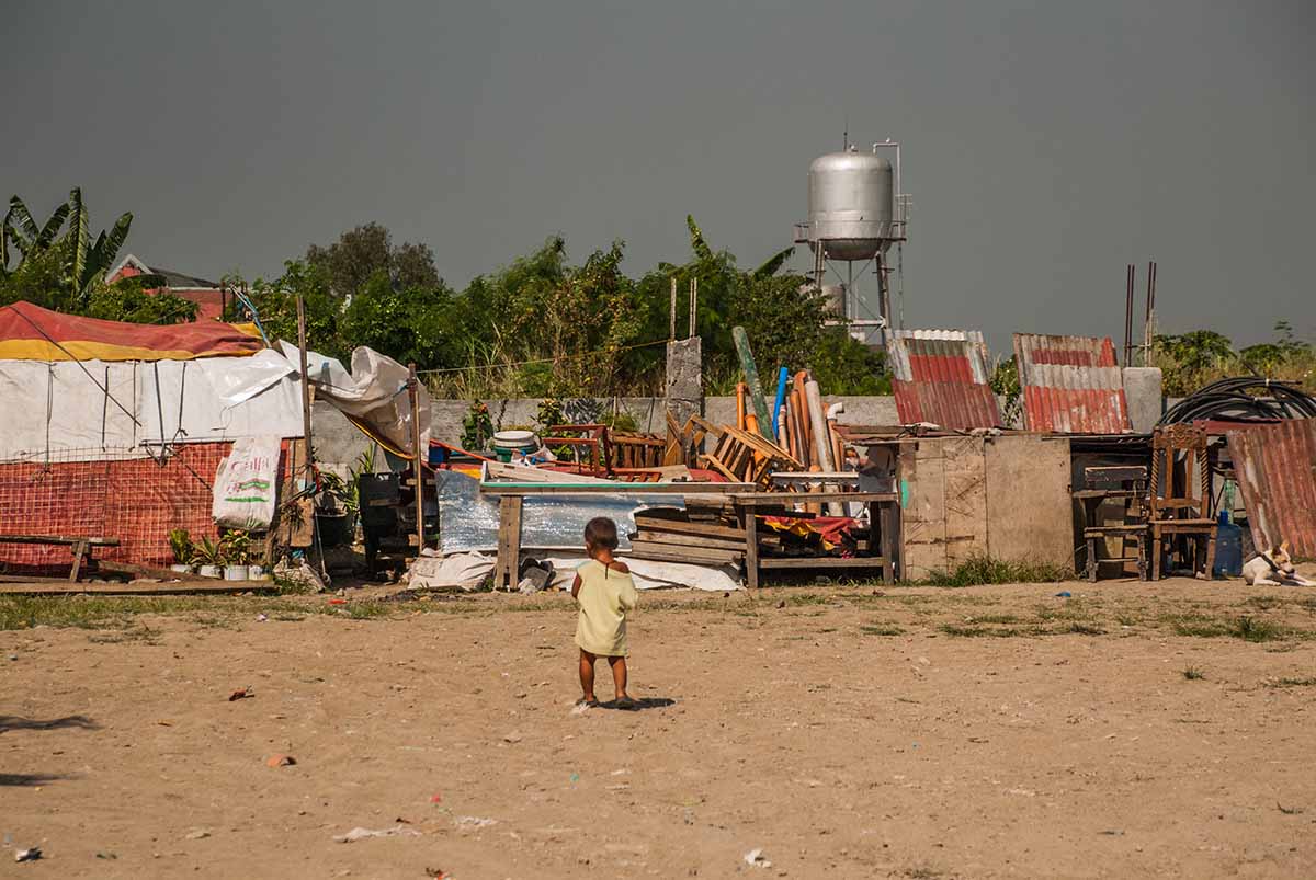 A child stands on an open dirt area with a refugee camp in the background A child stands on an open dirt area with a refugee camp in the background